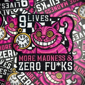 May include: A pink Cheshire Cat patch with the text "9 Lives" and "More Madness & Zero F*cks." The cat holds a gold pocket watch. The background features similar stickers.