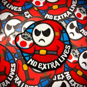 May include: A circular embroidered patch featuring a red-hooded character with a white mask and an angry expression. The character holds a red and white spotted mushroom. The text NO EXTRA LIVES is stitched in white along the bottom edge.