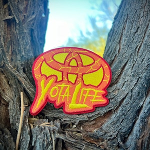 May include: A red and yellow embroidered patch with the text "Yota Life" and a stylized logo. The patch is attached to a tree trunk.