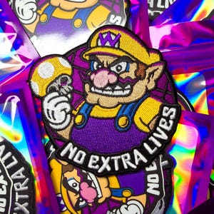 May include: Embroidered patch featuring a cartoon character in a yellow hat and purple overalls, holding a mushroom. The patch has the text "NO EXTRA LIVES" in white. The background is a holographic, iridescent material.