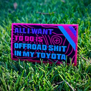 May include: Embroidered patch with the text "ALL I WANT TO DO IS OFFROAD SHIT IN MY TOYOTA" in blue, pink, and turquoise. The patch has a black background with a pink border and a design of lines. The patch is rectangular.