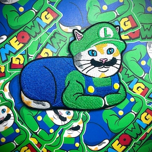 May include: A white cat wearing a green and blue Mario costume with a green hat and an 'L' on it. The cat is lying down with its paws crossed. The cat is surrounded by other patches with the words 'Meowig' on them.