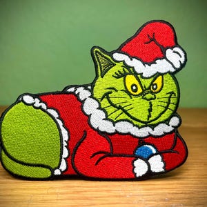 May include: Embroidered patch featuring a green cat-like character in a red Santa suit and hat with white trim. The character has a mischievous grin and is holding a blue ornament. The patch is on a wooden surface.