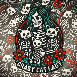 May include: Embroidered patch featuring a skeleton with teal hair and three skeleton cats. The patch is black with red and white accents, and the text "CRAZY CAT LADY" is included. The background is a pattern of cat skeletons and red flowers.