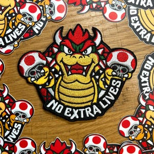 May include: Embroidered patch featuring Bowser from the Super Mario series, holding two mushroom-skull hybrids. The patch is red, yellow, and green with the text "NO EXTRA LIVES". Additional stickers with similar designs are scattered around.