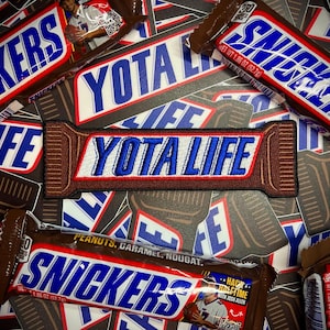 May include: An assortment of Snickers candy bars and patches. The patches read "YOTA LIFE" in blue and white lettering on a brown background. The candy bars have the word "SNICKERS" in blue and white lettering. The candy bars are 1.86 oz (52.7g).