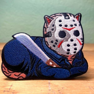 May include: Blue and white embroidered patch of a cat wearing a hockey mask and holding a knife.