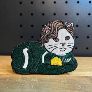 May include: Embroidered patch featuring a white cat with brown hair, wearing a green tracksuit with the number 456. The cat is holding a gold coin. The patch has a black background.