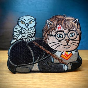 May include: Embroidered patch featuring a cat dressed as Harry Potter with a lightning bolt scar on its forehead, wearing glasses and a Gryffindor scarf, with a white owl perched on its back.