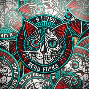 May include: A black and white embroidered patch of a cat with a skull face. The cat has red and turquoise details. The patch has the text "9 LIVES" and "ZERO FU*KS" around the cat.