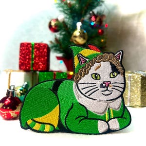 May include: A white cat wearing a green and yellow elf costume with a curly brown wig. The cat is lying down with its paws tucked under its body. The cat is in front of a Christmas tree and presents.