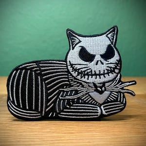 May include: Black and white embroidered patch of a cat with a stitched-up face, wearing a black and white striped suit. The cat is lying down with its head turned to the side.