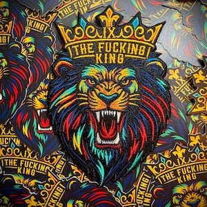 May include: Embroidered patch featuring a colorful lion's head wearing a crown. The text "THE FUCKING KING" is displayed above the lion's face. The lion's mane is a mix of blue, red, yellow, and green. The background is filled with similar patches and stickers.