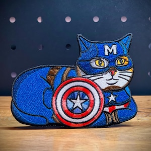 May include: A blue and red embroidered patch of a cat wearing a Captain America costume, including a red, white, and blue shield and a mask with the letter 'M' on it.
