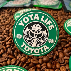 May include: Green and white patch with the text "Yota Life" and "Toyota" around a stylized coffee cup logo. The patch is surrounded by coffee beans.