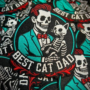 May include: Embroidered patch featuring a skeleton in a teal suit holding a skeleton cat. The patch has the words "BEST CAT DAD" in white lettering with red accents. The background is a pile of similar patches.