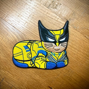 May include: A yellow and blue embroidered patch of a cat dressed as Wolverine, a superhero from Marvel Comics. The cat is wearing a yellow and blue costume with black claws and a black mask. The cat has a white mustache and a serious expression.