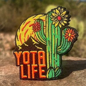 May include: Embroidered patch featuring a green cactus with yellow and red flowers, a yellow sun, and the text "YOTA LIFE" in orange letters. The patch has a black border and is set against a blurred desert background.