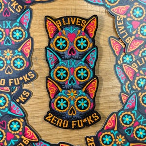 May include: A stack of three cat-shaped patches with sugar skull designs. The patches are black with colorful accents of teal, orange, pink, and yellow. The text "9 LIVES" and "ZERO FU*KS" are visible.