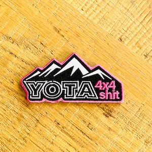 May include: Black and white embroidered patch with a pink outline. The patch features a mountain range with the text "YOTA 4x4 shit" in a bold font.