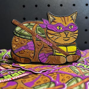 May include: Embroidered patch featuring a cat designed as a Teenage Mutant Ninja Turtle. The cat has a purple mask, a green shell, and a brown staff. The patch is on a pile of similar patches.
