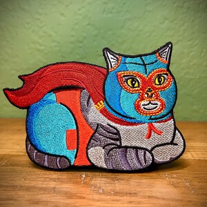 May include: Embroidered iron-on patch of a blue cat wearing a red and blue luchador mask. The cat is wearing a red cape and has a white and gray striped body.