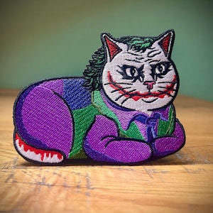 May include: Embroidered patch of a white cat with a purple, green, and blue costume. The cat has a Joker-inspired makeup design with a white face, red lips, and black eyes.