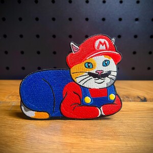 May include: A red, white, and blue embroidered patch of a cat wearing a Mario costume. The cat is lying down with its head turned to the side and has a mustache.
