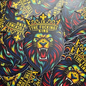 May include: A pile of stickers featuring a roaring lion wearing a crown. The lion's mane and face are a mix of vibrant colors, including red, yellow, and blue. The text "THE FUCKING KING" is printed above the lion's head.
