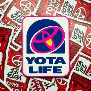 May include: A rectangular patch with a white background and a pink border. The design features a dark blue bell-shaped graphic containing a pink Toyota logo with a yellow center. Below the graphic, the text YOTA LIFE is printed in dark blue.
