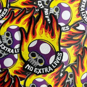 May include: Embroidered patch featuring a purple mushroom with white spots and a skull design, surrounded by flames in red, yellow, and black. The text "NO EXTRA LIVES" is displayed on a black banner. The background is filled with similar patches.