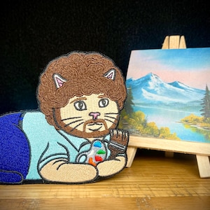 May include: Embroidered patch featuring a cat with brown fur and a beard, styled after a famous painter. The cat is holding a paintbrush and a paint palette. A miniature painting of a landscape is on a small wooden easel.