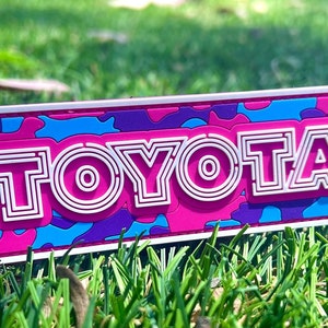 May include: A pink, blue, and purple camouflage patch with a white outline. The word "TOYOTA" is written in white letters with a pink outline.