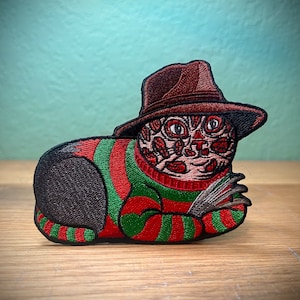 May include: Embroidered patch featuring a cat with a Freddy Krueger-inspired design. The cat wears a brown fedora and a red and green striped sweater. The cat's face has a scarred appearance, and it has metal claws.