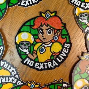 May include: Embroidered patch featuring Princess Daisy from the Super Mario series, holding a mushroom with a skull design. The patch is circular with a green grid background and the text "NO EXTRA LIVES". Additional patches are scattered around.