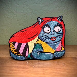 May include: A colorful embroidered patch of a cat with red hair, wearing a patchwork dress. The cat has blue eyes and a pink nose.