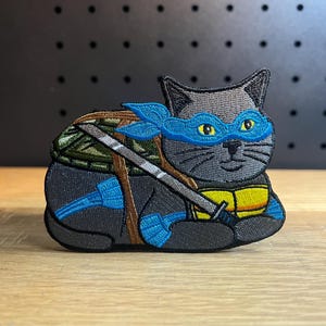May include: Embroidered patch featuring a gray cat in a blue mask and headband, resembling a ninja turtle. The cat has yellow eyes and a sword. The patch also includes a green and brown shell and blue accents.