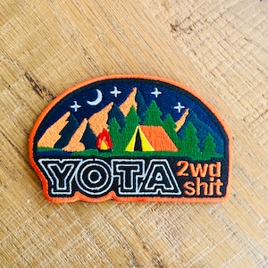May include: An orange and black embroidered patch with a camping scene, a crescent moon, stars, a campfire, a tent, and mountains. The text "YOTA 2wd shit" is in black and white.