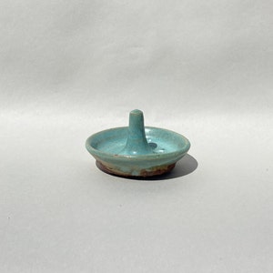 May include: A handmade ceramic ring holder in a light turquoise glaze. The ring holder has a circular base with a central post for holding rings. The base has a brown rim. The ring holder is on a light gray background.
