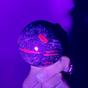Pode incluir: Um objeto esférico de pedra com uma superfície escura e salpicada, apresentando inclusões retangulares e lineares vermelhas vibrantes que brilham sob luz ultravioleta. A esfera possui uma textura lisa e polida com um reflexo distinto.