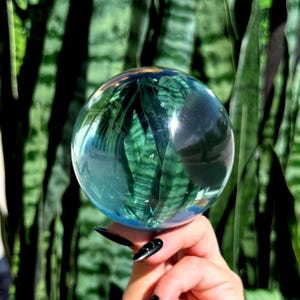 Könnte beinhalten: Eine klare, kugelförmige Glaskugel, die ihre Umgebung reflektiert. Die glatte, transparente Oberfläche der Kugel fängt eine verzerrte, umgekehrte Ansicht von grünen, länglichen, gestreiften Blättern ein und erzeugt einen visuellen Effekt von Tiefe.