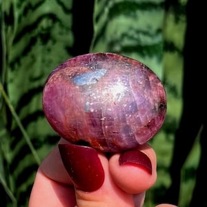 May include: A polished, oval-shaped ruby gemstone displaying deep magenta and purple hues with natural crystalline inclusions and reflective surfaces. The stone features a smooth, rounded texture with visible internal mineral variations.