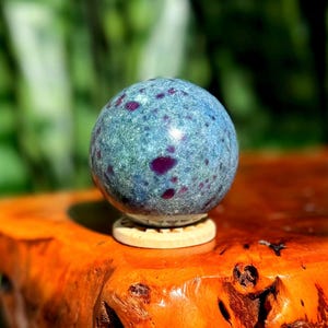 May include: A blue-green gemstone sphere with deep purple spots, resting on a small wooden stand. The sphere is polished and reflects light, showcasing its natural patterns. The background is a blurred green, with the sphere sitting on a wooden surface.