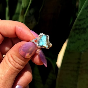 May include: A silver ring featuring a rectangular, iridescent blue gemstone. The ring's band has a unique open design on the sides. The gemstone displays shades of blue and green, set against a dark green background.