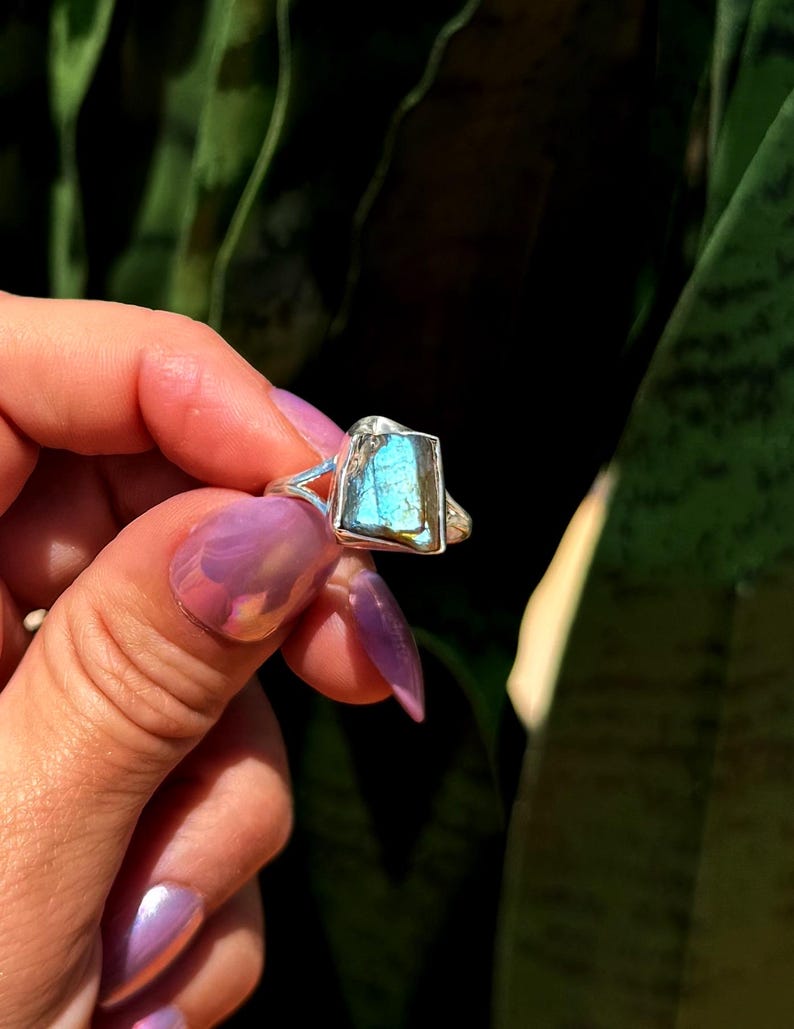 May include: A silver ring featuring a rectangular, iridescent blue gemstone. The ring's band has a unique open design on the sides. The gemstone displays shades of blue and green, set against a dark green background.