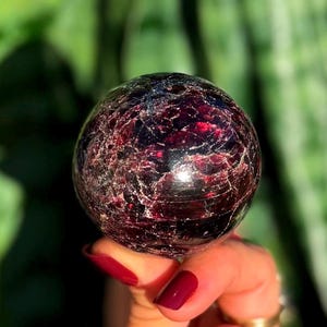 Puede incluir: Una gema esférica pulida, de color burdeos intenso, con intrincadas vetas blancas. La esfera se sostiene en una mano con esmalte de uñas burdeos a juego, sobre un fondo verde borroso.