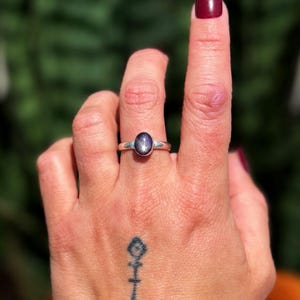 May include: A silver ring with an oval, dark blue gemstone. The ring is on a hand with a tattoo on the wrist and a painted fingernail. The background is a blurred green.