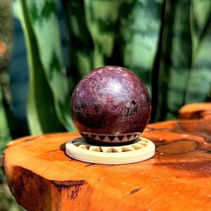 May include: A polished, deep burgundy sphere with dark inclusions and a distinct horizontal band of white triangular patterns near the base, resting securely on a circular, light-colored wooden stand with a sunburst-style carved edge.