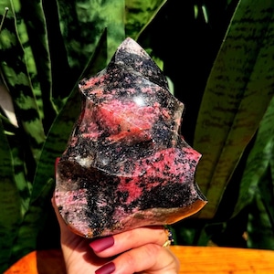 May include: A large, irregularly shaped, polished pink and black gemstone. The gemstone has a rough, natural texture and is a deep pink color with black veins running through it.