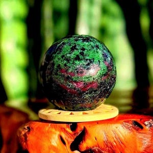 Peut inclure: Une pierre précieuse sphérique polie avec un mélange de couleurs vives. La sphère présente une surface verte tachetée, une bande rouge foncé et des touches de bleu et de noir. Elle repose sur un dessous de verre en bois clair, sur fond vert flou.